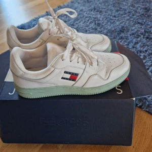 Tommy Jeans vita sneakers - Snygga vita sneakers från Tommy Jeans med klassisk logga i rött, blått och vitt på sidan. Skorna har snörning, perforerade detaljer på tån och en ljusgrön platt sula. Tillverkade i skinn och syntetmaterial för en fräsch och modern look.