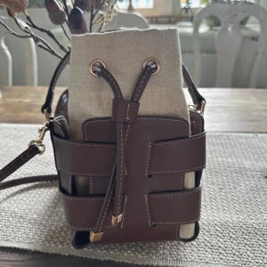 Bucket bag hm  - Använd 2 gånger!!! Snygg axelväska från H&M med cool bucket-form. Väskan har bruna detaljer i skinn och beige tyg, samt snörning upptill. Guldiga metalldetaljer och justerbar axelrem gör den både praktisk och trendig. Perfekt storlek för mobil, plånbok och småsaker.💞