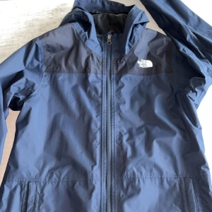 Blå vindjacka från The North Face  - Snygg blå vindjacka från The North Face med huva och hel dragkedja framtill. Jackan har mörkare partier över axlarna och klassisk logga på bröstet. Perfekt för blåsiga dagar och enkel att matcha med streetwear. Materialet är lätt och känns sportigt. Pris går o diskutera 