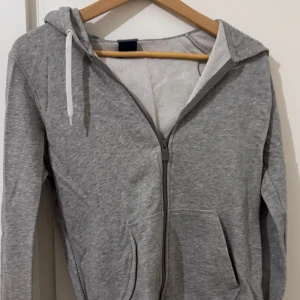 Grå hoodie från Nike med dragkedja - Säljer en grå hoodie från Nike i storlek M. Tröjan har huva med vita snören, hel dragkedja framtill och två fickor. Nike-loggan är broderad på bröstet. Perfekt för chill eller träning, tillverkad i mjuk bomullsmix för skön känsla.