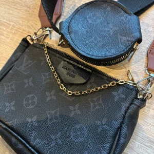  svart axelväska med kedja - Snygg svart axelväska  med klassiskt monogrammönster och guldfärgad kedja. Väskan har bred axelrem med bruna detaljer och en extra rund liten ficka framtill. Perfekt för dig som vill ha en trendig och praktisk väska med lyxig känsla.