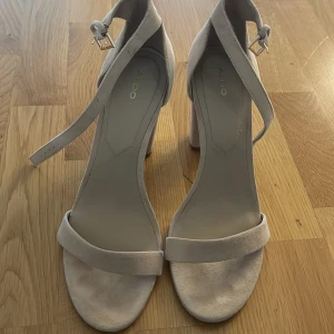 Beige klackade sandaler från ALDO - Stilrena sandaler från ALDO i beige mocka med bred klack och öppen tå. Skorna har en enkel rem över tårna och justerbar ankelrem med spänne. Perfekta för dig som gillar en clean och modern look med lite höjd.