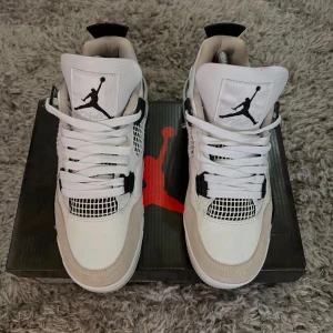  Jordan 4 vit/svart/grå - Nike Air Jordan 4 sneakers i vitt, svart och ljusgrått. Skorna har klassisk meshpanel, snörning och Jumpman-logga på plös och häl. Ovandel i skinn och mocka med svarta detaljer och vit sula. Perfekt för dig som vill sticka ut med en ikonisk siluett.