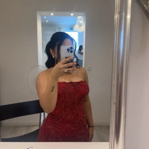 Röd glittrig axelbandslös balklänning - Säljer en röd långklänning med glittriga paljettdetaljer och vackert mönster. Klänningen är axelbandslös och har en figurnära passform som framhäver siluetten. Perfekt för bal eller fest. Materialet känns lyxigt och skimrar snyggt i ljuset.