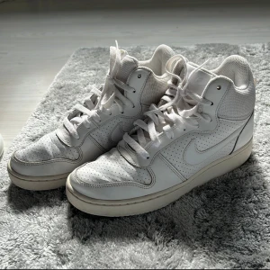 Nike Air Force 1 vita sneakers - Säljer ett par klassiska Nike Air Force 1 i helvitt skinn med perforerade detaljer och högt skaft. Skorna har snörning och en platt sula. Perfekta för dig som gillar en clean och tidlös look. Storlek 39 (US 8).