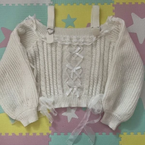 Vit stickad croppad topp med rosetter - Supersöt vit stickad croppad topp med breda axelband, spetskant och dekorativa rosetter framtill. Ballongärmar med transparenta band längs sidorna och hjärtformade detaljer på banden. Perfekt för dig som gillar gulliga och unika plagg.