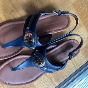 Blå sandaler från Tommy Hilfiger - Snygga sandaler från Tommy Hilfiger i marinblått skinn med bruna sulor. Dekorativa metall-loggor framtill och justerbar rem runt hälen. Perfekta för sommaren och ger en stilren touch till din outfit.