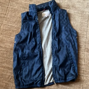 Mörkblå väst från Gucci med GG-mönster - Snygg mörkblå väst från Gucci med ikoniskt GG-mönster över hela tyget. Västen har dragkedja framtill, ståkrage och grått innerfoder med en praktisk innerficka. Perfekt för dig som vill ha en exklusiv och stilren look. Finns en luva att fälla ut! Den är för tolv och 13 år, en jävligt fet väst. Har påse till om man vill ha den är och Gucci!