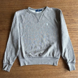 Grå sweatshirt från Polo Ralph Lauren - Snygg grå sweatshirt från Polo Ralph Lauren med klassisk rund halsringning och broderad vit logga på bröstet. Tröjan har långa ärmar och ribbade muddar vid ärmslut och nederkant. Perfekt för en chill och stilren look. Passar 11-13 år 