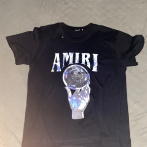 Svart Amiri t-shirt med tryck - Snygg svart t-shirt från Amiri med stort tryck framtill av en hand som håller en glaskula och AMIRI-loggan ovanför. T-shirten har klassisk passform och korta ärmar. Perfekt för dig som vill sticka ut med en cool streetstyle-look.