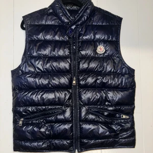 Mörkblå dunväst från Moncler - Snygg mörkblå dunväst från Moncler med glansig finish och klassisk logga på bröstet. Västen har hög krage, två dragkedjefickor framtill och stängs med dragkedja. Perfekt för lager-på-lager och ger en sportig vibe.