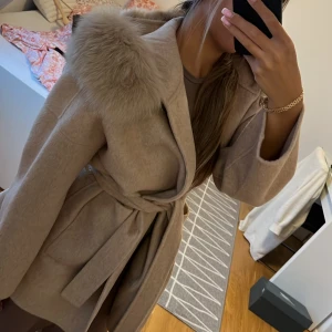 Beige kappa med pälsdetaljer och bälte - Snygg beige kappa med stora pälsdetaljer på kragen och knytskärp i midjan. Jackan har en loose passform och är perfekt för kyligare dagar. Materialet känns mjukt och lyxigt, och den har långa ärmar för extra värme.