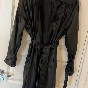Svart trenchcoat i skinnimitation - Säljer en svart trenchcoat i skinnimitation med bälte i midjan och stora slag. Jackan har långa ärmar med spänndetaljer vid handlederna och knappar framtill. Perfekt för dig som vill ha en edgy och stilren look.