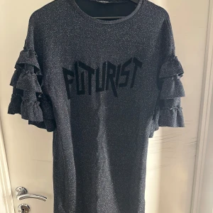 Glittrig svart kortklänning med volangärm - Cool svart kortklänning med glittrigt tyg och texten 'FUTURIST' framtill. Klänningen har korta ärmar med flera volanger som ger en unik och trendig look. Perfekt för dig som gillar att sticka ut och vill ha något extra till garderoben.