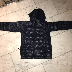 Mörkblå dunjacka från Peak Performance - Säljer en mörkblå dunjacka från Peak Performance med huva och dragkedja framtill. Jackan har quiltad design, två fickor med dragkedja och lång ärm. Perfekt för kyliga dagar och enkel att matcha med olika outfits. Jackan är i dåligt skick NYPRIS:2000 Säljer jackan för 350
