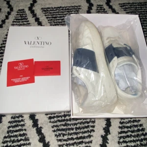 Valentino Garavani Open sneakers vit/blå - Säljer ett par Valentino Garavani Open sneakers i vitt med bred blå rem över sidan. Skorna har en chunky platt sula och rund tå, tillverkade i skinn. Kommer med originalkartong och dustbag. Perfekt för dig som gillar stilrena designer och vill sticka ut med exklusiva sneakers. Skorna ligger på storlek 43 och kan tänka mig att sänka att sänka på snabbäffär.