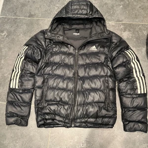 Adidas jacka  - Svart pufferjacka från Adidas med klassiska vita ränder på ärmarna och logga på bröstet. Jackan har huva, dragkedja framtill och två sidofickor med dragkedja. Perfekt för kalla dagar och riktigt snygg streetstil.