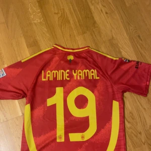 Spanien Lamine Yamal #19 EM 2024 tröja - Säljer en officiell Spanien fotbollströja från EM 2024 med Lamine Yamal och nummer 19 på ryggen. Tröjan är röd med gula detaljer, Adidas-logga, landslagsemblem och turneringspatch på ärmarna. Tillverkad i lätt och ventilerande material, perfekt för match eller träning.