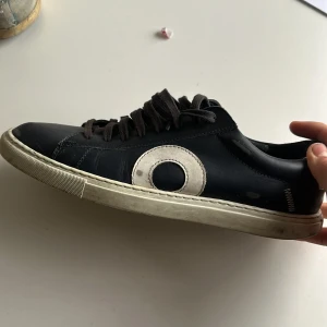 Svarta sneakers från Oliver Cabell - Snygga svarta sneakers från Oliver Cabell med vit rund detalj på sidan och vit häl. Skorna har platt sula, rund tå och svarta snören. Den ena har inga sko snören för att dom gick sönder så man får själv lösa nya till dom.