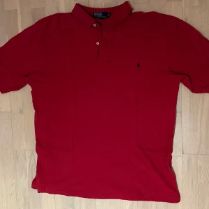 Röd pikétröja från Ralph Lauren - Klassisk röd pikétröja från Ralph Lauren med korta ärmar och krage. Tröjan har knäppning med två knappar och den ikoniska lilla broderade loggan på bröstet. Tillverkad i mjuk bomull som känns skön mot huden. Perfekt för en stilren och avslappnad look.