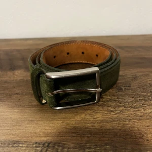 Grönt mockabälte från Oscar Jacobson - Snyggt bälte i mörkgrön mocka från Oscar Jacobson med klassiskt silverfärgat spänne. Insidan är i ljust skinn med synliga sömmar. Perfekt accessoar för att lyfta din outfit med en stilren touch.