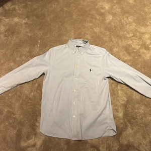 skjorta från  Ralph Lauren - Klassisk ljusblå skjorta från Polo Ralph Lauren i slim fit-modell. Skjortan har långa ärmar, knappar framtill och ikonisk broderad logga på bröstet. Tillverkad i mjuk bomull och passar perfekt till en stilren look. Den är storlek Xs/S, Priset är 449. Nypris ligger på ungefär 2000, priset är diskuterbart. Skriv vid vilken fråga som helst!