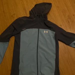 Under Armour vindjacka med huva M - Använd 2 gånger, den är som ny. vindjacka från Under Armour i mörkblå och ljusblått. Jackan har huva, hel dragkedja och logga på bröstet samt stort tryck på ryggen. Perfekt för dig som gillar sportig stil och vill ha något lätt och funktionellt.