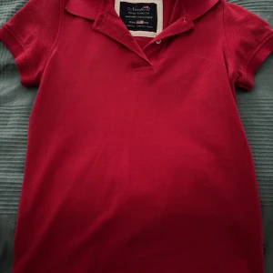 Röd polo t-shirt från Newport XS - Snygg röd polo t-shirt från Newport i storlek XS. Klassisk pikékrage och korta ärmar, tillverkad i bomull med en clean look. Perfekt för dig som gillar preppy stil och vill ha en enkel men stilren t-shirt med amerikansk vibe.