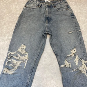 Blå ripped jeans med loose fit - Säljer ett par blå jeans från Zara med rejält slitna och trasiga detaljer på båda benen. Jeansen har en loose fit och klassisk femficksdesign. Perfekta för dig som gillar en avslappnad och edgy stil. Materialet är klassisk denim i bomull.