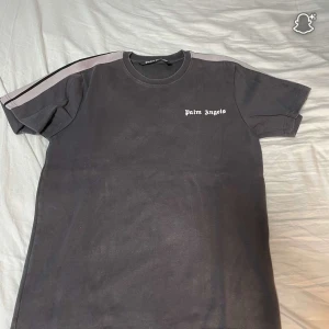 Grå Palm Angels t-shirt med ränder - Palm Angels t-shirt i grå med vita ränder längs axlarna och logotyp tryckt på bröstet. Klassisk rund hals och korta ärmar. T-shirten har en loose passform och är gjord i mjuk bomull, perfekt för en chill streetwear-look.