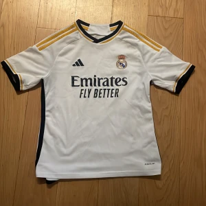 Real Madrid hemmatröja Adidas 13-14Y - Säljer en officiell Real Madrid hemmatröja från Adidas i storlek 13-14 år. Tröjan är vit med svarta och gula detaljer, klubbmärke och Adidas-logga på bröstet samt 'Emirates Fly Better' tryck. Tillverkad i lätt och ventilerande polyester med korta ärmar.