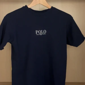 Marinblå t-shirt från Polo Ralph Lauren - Snygg marinblå t-shirt från Polo Ralph Lauren med klassisk vit logga broderad på bröstet. T-shirten är kortärmad och har en normal passform. Perfekt till jeans eller shorts för en clean och stilren look.