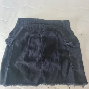 Svart jeanskjol med rå kant - Cool svart jeanskjol med rå, fransig kant nertill och praktiska fickor framtill. Kjolen har bälteshällor och stängs med dragkedja och knapp. Perfekt för dig som gillar en avslappnad och trendig stil.