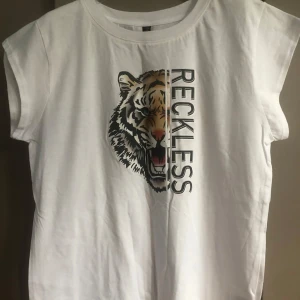 Vit t-shirt PREPAIR med tigertryck - Vit t-shirt från PREPAIR i 100% bomull med coolt tigertryck och texten 'RECKLESS' framtill. T-shirten har korta ärmar och en avslappnad passform. Perfekt för dig som vill sticka ut med en edgy look.