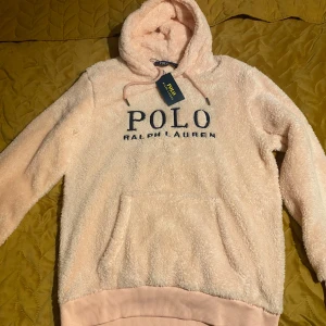 Ljusrosa fleece hoodie Polo Ralph Lauren - Supermjuk och fluffig ljusrosa hoodie från Polo Ralph Lauren med stor broderad logga framtill. Tröjan har huva med snörning, magficka och ribbade muddar. Perfekt för dig som vill ha en cozy och snygg look under kyliga dagar.