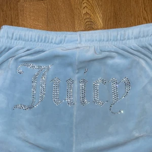 Ljusblå Juicy Couture mjukisbyxor - Supermysiga ljusblå mjukisbyxor från Juicy Couture i velour med strassdetaljer och snörning i midjan. Byxorna har resår vid benslut och midja samt glittrande Juicy-text på baksidan och JC-logga fram. Perfekta för en chill och trendig look.