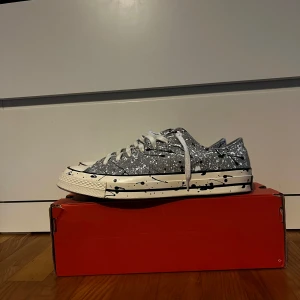 Grå Converse sneakers med stänkmönster - Säljer ett par grå Converse sneakers med vit sula och coolt stänkmönster i vitt och svart. Skorna har klassisk rund tå, vita snören och platt sula. Perfekta för dig som vill sticka ut med en unik look.