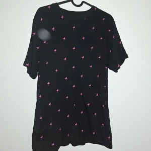 Svart t-shirt med rosa flamingos FSBN - Svart t-shirt från FSBN i storlek S med små rosa flamingos som mönster över hela plagget. Modellen är regular fit med rund hals och korta ärmar. Materialet är mjuk bomull som känns skönt mot huden. Perfekt för dig som gillar coola prints och vill sticka ut lite extra.