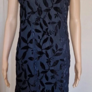 Marinblå långklänning Woman Collection - Snygg marinblå långklänning från Woman Collection i storlek 34. Klänningen har ett sammetsaktigt utstående blommigt mönster, v-ringad hals och är ärmlös. Materialet känns lyxigt och har en lätt transparent känsla nedtill. Perfekt för dig som vill sticka ut med stilren elegans.