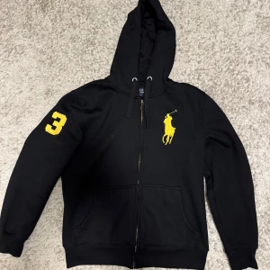 Svart kofra från Polo Ralph Lauren - Svart kofta från Polo Ralph Lauren med gul logga på bröstet och en gul siffra 3 på ärmen. Tröjan har dragkedja, huva med snörning och känguruficka. Perfekt för dig som gillar sportig och stilren. Priset kan också diskuteras vid snabb affär! ☺️