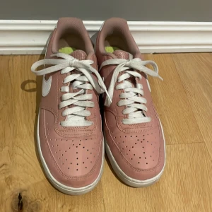 Nike sneakers i rosa och vitt - Säljer ett par snygga Nike sneakers i rosa med vita detaljer och vit sula. Skorna har klassisk snörning, rund tå och är tillverkade i syntetmaterial med skinnkänsla. Använda ett fåtal gånger.