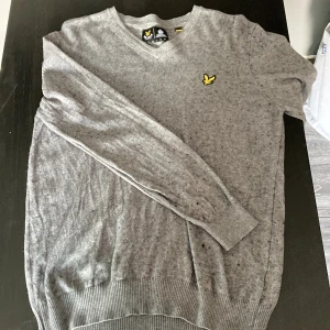Grå stickad tröja från Lyle & Scott - Snygg grå stickad tröja från Lyle & Scott med klassisk v-ringning och den ikoniska gula örnloggan broderad på bröstet. Tröjan har långa ärmar och ribbade muddar för en clean look. Perfekt för lager-på-lager och chill stil.