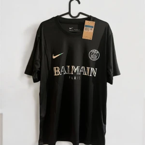 Svart PSG x Balmain fotbollströja Nike - Snygg svart fotbollströja från Nike i samarbete med PSG och Balmain Paris. Tröjan har korta ärmar, rund hals och tryck med Balmain-logga samt PSG-emblem på bröstet. Materialet är lätt och andas, perfekt för träning eller match.