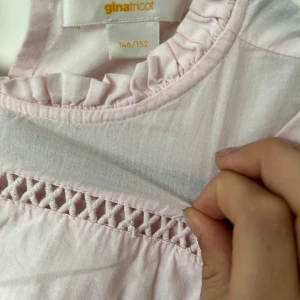 Ljusrosa blus med volang från Gina Tricot - Supersöt ljusrosa blus från Gina Tricot med volangdetaljer vid axlar och hals, samt ett broderat parti över bröstet. Blusen har korta puffärmar och är tillverkad i mjuk bomull. Perfekt för en söt och avslappnad look.