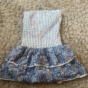 Vit och blå axelbandslös kortklänning - Supersöt axelbandslös kortklänning i vitt och blått med smockad överdel och volangkjol. Klänningen har ett grafiskt mönster. Perfekt för varma dagar och har en fräsch, ungdomlig vibe med fina detaljer längs kanterna. Från Gina tricot 