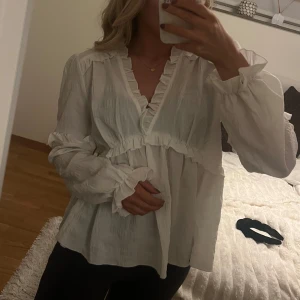 Vit volangblus med v-ringning nakd - Supersöt vit blus med v-ringning och volangdetaljer längs halsringning, ärmslut och över bysten. Lätt och luftig känsla med långa ärmar och avslappnad passform🤩använd endast 2 gånger så den är som ny! Eftersom den är så luftig passar den även en 38a