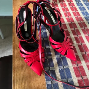 Röda pumps med snörning från Zara - Säljer ett par röda pumps från Zara med spetsig tå och snygga remmar som går över foten och runt ankeln. Klackarna har en elegant design och är tillverkade i blankt syntetmaterial. Perfekt för dig som vill sticka ut med färg och coola detaljer.