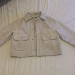 Beige overshirt från ZARA XS - Snygg beige overshirt från ZARA i storlek XS. Jackan har stora fickor framtill med tryckknappar, klassisk krage och raka ärmar. Materialet är mjukt och ger en clean look, perfekt att slänga över en topp. Enkel och stilren design.