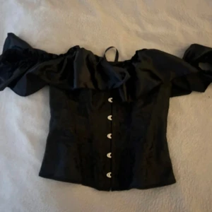 Svart offshoulder korsett med snörning - Säljer en svart korsett med offshoulder och puffiga volangärmar. Toppen har snygg snörning i ryggen och knappar framtill. Materialet är glansigt och har ett diskret mönster. Perfekt till halloween! Aldrig använt!
