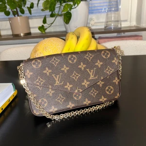 HM axelväska - Säljer en brun axelväska från HM med klassiskt monogrammönster och guldfärgad kedja. Väskan har kuvertformad front och vinrött innerfoder. Perfekt storlek för det viktigaste och ger en lyxig touch till din outfit. Ps har tappat bort gulddetaljen på framsidan. 